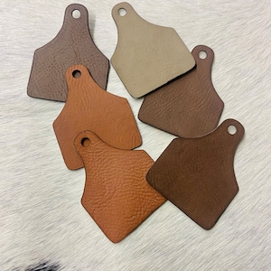 May include: Six leather ear tags in various shades of brown, including tan, light brown, dark brown, and a reddish brown. The tags are shaped like a traditional ear tag with a hole at the top.