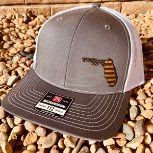 May include: A gray and white trucker hat with a brown and black Florida state outline with an American flag design. The hat has a white mesh back and a black brim. The hat has a label that says "Richardson 112 Original Trucker Adjustable".