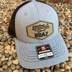 Pode incluir: Bon&eacute; de caminhoneiro juvenil cinza e preto com um remendo marrom que apresenta um peixe perca e a palavra "BOAZ". O bon&eacute; tem uma parte de tr&aacute;s de malha preta e uma etiqueta preta "Richardson" com o texto "EST. 1970 ORIGINAL 112 TRUCKER YOUTH".