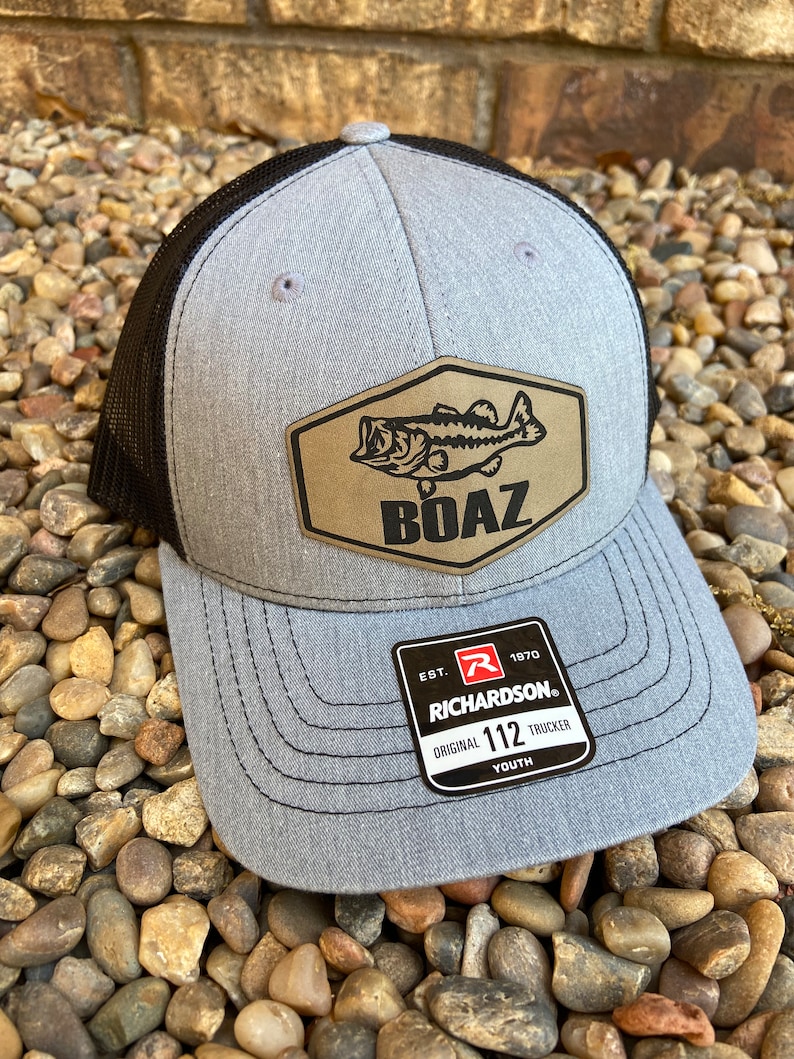 Pode incluir: Bon&eacute; de caminhoneiro juvenil cinza e preto com um remendo marrom que apresenta um peixe perca e a palavra "BOAZ". O bon&eacute; tem uma parte de tr&aacute;s de malha preta e uma etiqueta preta "Richardson" com o texto "EST. 1970 ORIGINAL 112 TRUCKER YOUTH".