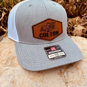 May include: A gray and white trucker hat with a brown leather patch that says "COLTON" and features a welder with an American flag. The hat is adjustable and has a Richardson 112 label.