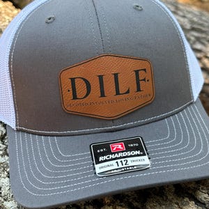 May include: A gray and white trucker hat with a brown leather patch that says "DILF" in black letters. The patch also says "Dedicated Involved Loving Father". The hat has a white mesh back and a black adjustable strap with a black label that says "Richardson 112 Original Trucker Adjustable".