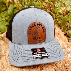 May include: A gray and black trucker hat with a brown leather patch featuring a bird and the text "Little Pecker Club, Little Gents Making Dents". The hat also has a red and white label with the text "Est. 1970 Richardson Original 112 Trucker Adjustable".