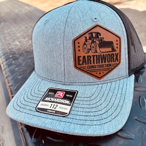 May include: A blue and black trucker hat with a brown leather patch featuring the text "EARTHWORX CONSTRUCTION" and an image of a bulldozer. The hat has a curved brim and a mesh back. The tag reads "RICHARDSON 112 TRUCKER YOUTH".