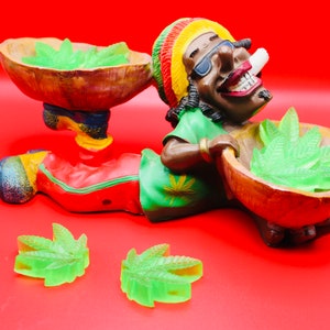 May include: A green and brown ceramic figurine of a person wearing a red, yellow, and green hat and holding two bowls filled with green leaf-shaped objects.