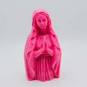 May include: A pink figurine of the Virgin Mary with her hands clasped in prayer. She is wearing a long, flowing robe and a veil.