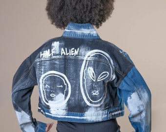 May include: A cropped denim jacket with a black and blue wash. The back of the jacket features white painted alien-themed illustrations and the words "HALF ALIEN". The jacket has long sleeves and a button-up front.