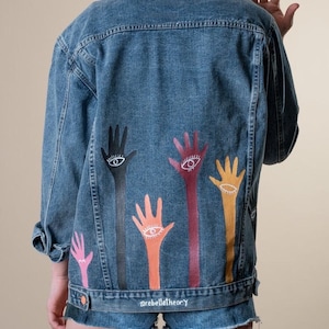 May include: A blue denim jacket featuring hand-painted eye designs in various colours. The jacket has a button-up front and long sleeves. The text "@rebelletheory" is printed at the bottom. A stylish, artistic piece.