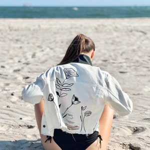 Könnte beinhalten: Weiße Jeansjacke mit schwarzem Blumendesign auf dem Rücken. Die Jacke hat einen ausgefransten Saum und einen schwarzen Kragen. Die Person sitzt an einem Sandstrand mit dem Meer im Hintergrund.