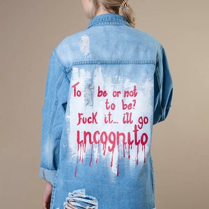 Könnte beinhalten: Hellblaue Jeansjacke mit Distressed-Details. Auf der Rückseite der Jacke steht der Text "To be or not to be? Fuck it... I'll go incognito" in roter und weißer Farbe. Aufgerollte Ärmel.