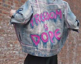 May include: A light blue denim jacket with a pink collar and the words "FREEDOM IS DOPE" in pink graffiti-style lettering on the back. The jacket has a distressed look.