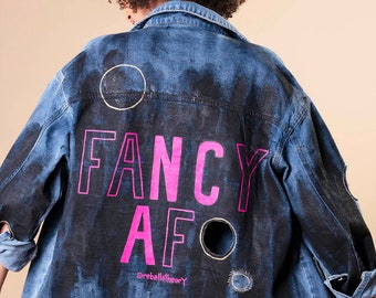 May include: Blue denim jacket with black paint effect and distressed details. The back features the words "FANCY AF" in bright pink, with the @rebeltheory logo. A unique, artistic fashion statement.