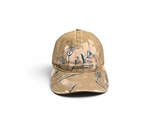 May include: A tan baseball cap with a distressed, washed-out finish featuring dark blue line art of stylized flowers and leaves. The design includes thin, hand-drawn botanical illustrations across the front panels and the curved brim.