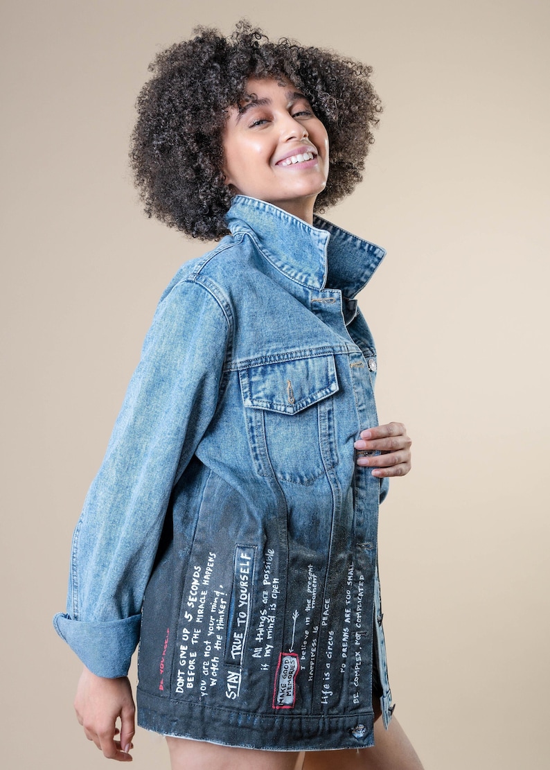 K&ouml;nnte beinhalten: Hellblaue Jeansjacke mit hochgekrempelten &Auml;rmeln. Die Jacke hat ein dunkelgraues Design auf dem unteren R&uuml;cken mit wei&szlig;em Text, darunter der Satz &bdquo;DON'T GIVE UP 5 SECONDS BEFORE THE MIRACLE HAPPENS&ldquo; und andere inspirierende Zitate. Klassischer Stil.