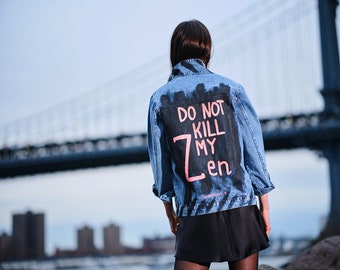 Può includere: Giacca di jeans blu con le parole "DO NOT KILL MY ZEN" in lettere rosa e bianche. La giacca ha un design nero dello skyline. Lo sfondo include un ponte e degli edifici.