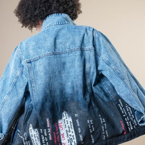 K&ouml;nnte beinhalten: Hellblaue Jeansjacke mit verblasstem schwarzem R&uuml;cken. Auf der R&uuml;ckseite der Jacke befindet sich wei&szlig;er Text mit inspirierenden Zitaten und Phrasen. Die Jacke hat einen klassischen Kragen und eine Knopfleiste.