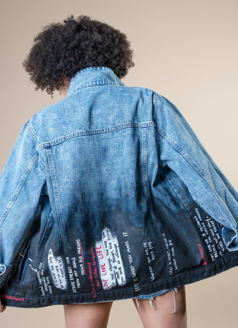 K&ouml;nnte beinhalten: Hellblaue Jeansjacke mit verblasstem schwarzem R&uuml;cken. Auf der R&uuml;ckseite der Jacke befindet sich wei&szlig;er Text mit inspirierenden Zitaten und Phrasen. Die Jacke hat einen klassischen Kragen und eine Knopfleiste.