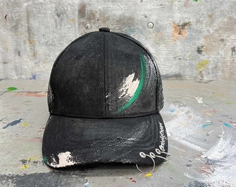 May include: Black baseball cap with white and green paint strokes. The brim has a silver ring and the text "@cirebelletheory". The cap is a stylish accessory.