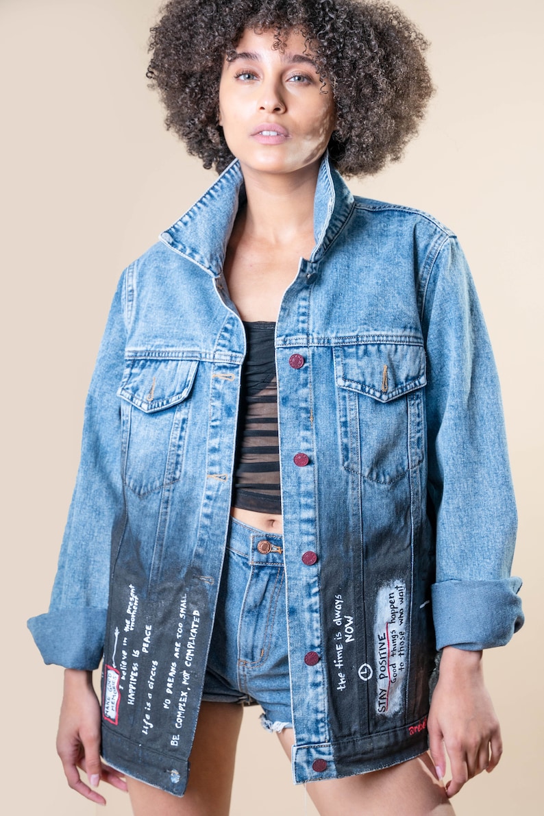 K&ouml;nnte beinhalten: Hellblaue Jeansjacke mit verblasstem schwarzem Unterteil, handschriftlichem Text und roten Kn&ouml;pfen. Die Jacke wird mit Jeansshorts kombiniert. Der Text enth&auml;lt S&auml;tze wie "Stay Positive" und "The time is always NOW."