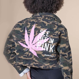 Könnte beinhalten: Camouflage-Jeansjacke mit einem rosa Cannabisblatt und dem Schriftzug "SEE LIFE IN PINK" auf dem Rücken. Die Jacke hat eine verkürzte Passform und hochgekrempelte Ärmel. Der Markenname "@rebeltheory" ist ebenfalls aufgedruckt.