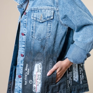 K&ouml;nnte beinhalten: Hellblaue Jeansjacke mit verblasstem schwarzem Unterteil. Die Jacke hat handgeschriebenen Text, darunter "STAY POSITIVE" und "Good things happen to those who wait". Sie hat rote Kn&ouml;pfe und einen klassischen Kragen.