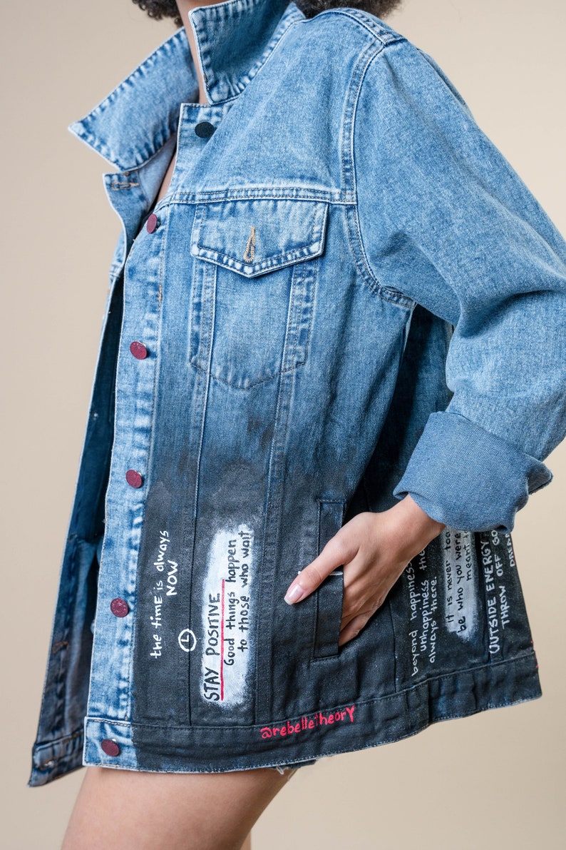 K&ouml;nnte beinhalten: Hellblaue Jeansjacke mit verblasstem schwarzem Unterteil. Die Jacke hat handgeschriebenen Text, darunter "STAY POSITIVE" und "Good things happen to those who wait". Sie hat rote Kn&ouml;pfe und einen klassischen Kragen.