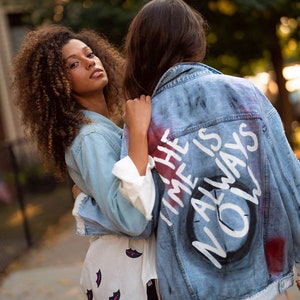 Könnte beinhalten: Zwei Personen umarmen sich. Eine trägt eine hellblaue Jeansjacke mit dem Aufdruck "THE TIME IS ALWAYS NOW" auf dem Rücken. Die andere trägt ein weißes Hemd mit einem schwarzen Lippenmuster.