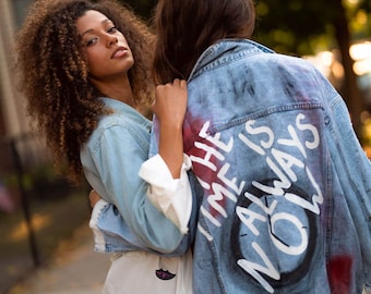 May include: Two people are embracing, one wearing a light blue denim jacket with the words "THE TIME IS ALWAYS NOW" painted on the back. The other person is wearing a white shirt with a black lip pattern.