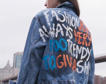 May include: A blue denim jacket with white, orange, and blue graffiti-style text on the back. The text reads "FASHION ALWAYS TOO TRENDY TO GIVE A SH*T". The jacket has a classic collar and buttoned cuffs.