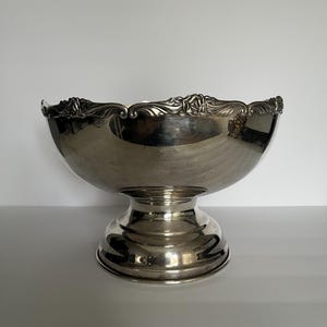 May include: A polished silver-toned pedestal bowl featuring a wide, rounded basin and a sturdy, circular base. The upper rim is decorated with a detailed, embossed floral pattern that encircles the entire circumference of the bowl.