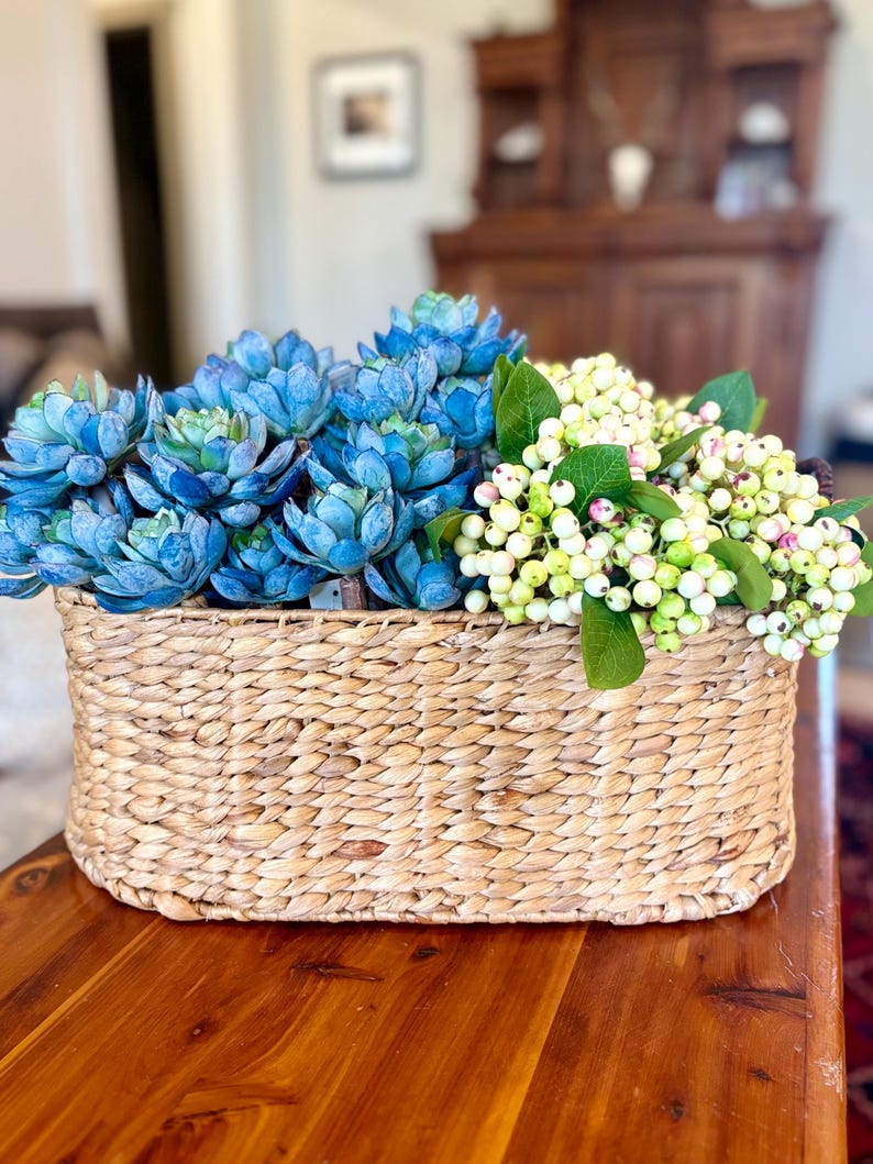 May include: A rectangular woven basket overflowing with blue succulents and white berry stems. The basket is made from natural fibres. The succulents display a gradient of blue shades, and the berries are accented with green leaves.
