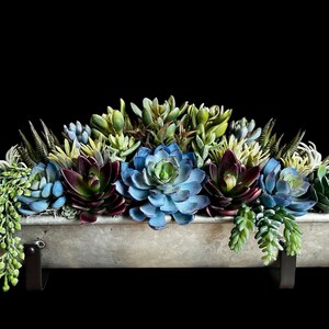 May include: A metal trough planter filled with artificial succulents in shades of blue, green, and burgundy. The planter is suspended by black metal brackets. The background is dark. The planter is approximately 60 cm long.