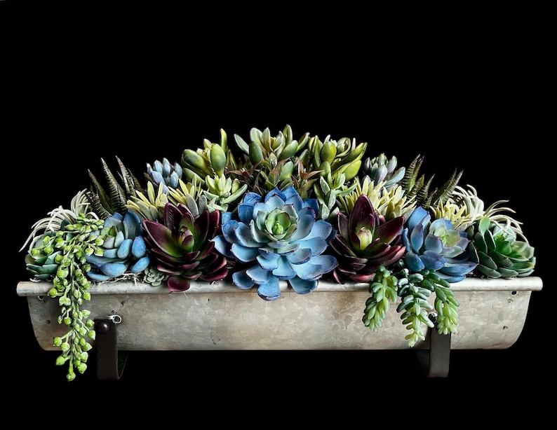May include: A metal trough planter filled with artificial succulents in shades of blue, green, and burgundy. The planter is suspended by black metal brackets. The background is dark. The planter is approximately 60 cm long.