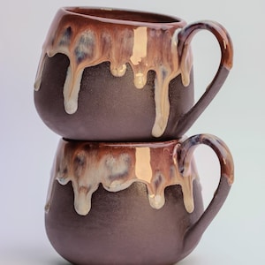 May include: Two stacked ceramic mugs. The dark brown mugs have a distinctive glaze with a dripped effect in shades of brown, beige, and white. Each mug has a curved handle. Ideal for tea or coffee, these mugs are a stylish addition to any kitchen.
