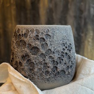 May include: A gray ceramic planter with a textured surface that resembles the moon. The planter is sitting on a white fabric surface.