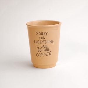 May include: A beige ceramic coffee cup with the text "SORRY FOR EVERYTHING I SAID BEFORE COFFEE" in black handwritten font. The cup has a slightly flared rim and a tapered body, perfect for a morning beverage.