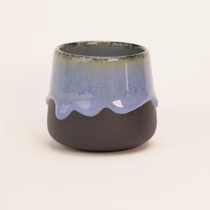 Pode incluir: Uma caneca de cerâmica preta com um esmalte azul claro que escorre pelas laterais. O esmalte tem um brilho iridescente sutil.