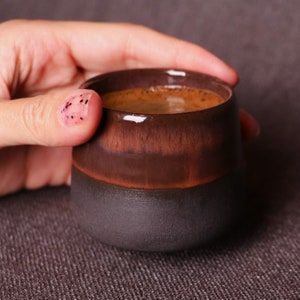 May include: A close-up of a dark brown and black ceramic mug filled with coffee. The mug is being held by a hand with pink nail polish.