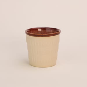 Könnte beinhalten: Eine weiße Keramiktasse mit braunem Rand. Die Tasse hat ein strukturiertes, geripptes Design.