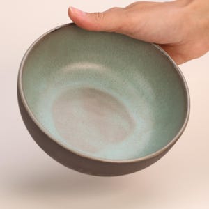 Può includere: Una ciotola in ceramica verde con un bordo marrone scuro. La ciotola è vuota e ha una glassa macchiata.