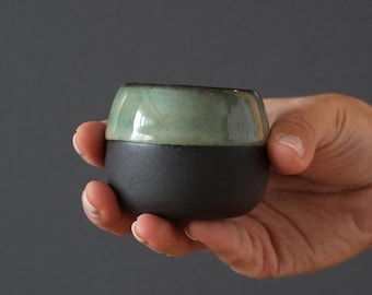 May include: A small, round ceramic cup with a black base and a green glaze on the top half.