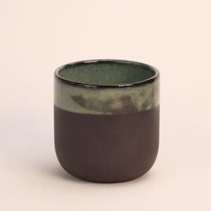 Pode incluir: Uma caneca de cerâmica marrom escura com um esmalte verde e marrom. A caneca tem um fundo arredondado e uma borda larga.