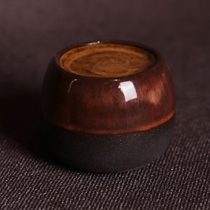 Puede incluir: Una taza pequeña, redonda, de cerámica marrón y negra con un líquido marrón en su interior. La taza tiene un acabado brillante y un ligero borde alrededor del borde.