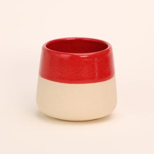 Op de afbeelding: Een rode en witte keramische mok met een afgeronde bodem. De mok is geglazuurd in een effen rode kleur op de bovenste helft en een effen witte kleur op de onderste helft.