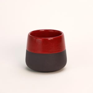 Op de afbeelding: Een rode en zwarte keramische mok met een afgeronde bodem. De bovenste helft van de mok is rood en de onderste helft is zwart.