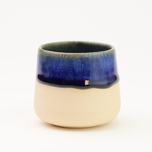 Peut inclure: Un gobelet en céramique fait à la main avec un design bicolore. La moitié supérieure est un émail bleu foncé, passant à une couleur crème plus claire en bas. La tasse a une forme arrondie et légèrement irrégulière.