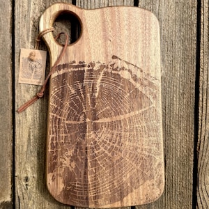 May include: Wooden cutting board with a handle and a tree ring design. The board is rectangular with rounded edges and a hole in the handle. A small tag is attached with a leather cord. The wood has a natural grain and color.