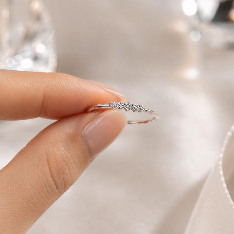 May include: A delicate silver ring featuring five small, sparkling diamonds set in a row. The ring is held between fingers, showcasing its minimalist design and elegant simplicity. The diamonds catch the light, creating a subtle sparkle.