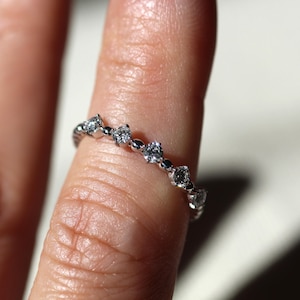May include: A delicate silver ring with a repeating pattern of small, sparkling diamonds. The ring is set with multiple round and marquise-cut stones, creating a continuous band of light. The ring is shown on a finger.