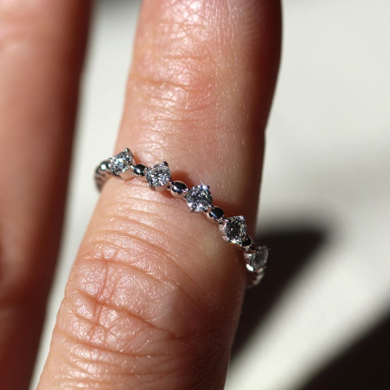 May include: A delicate silver ring with a repeating pattern of small, sparkling diamonds. The ring is set with multiple round and marquise-cut stones, creating a continuous band of light. The ring is shown on a finger.